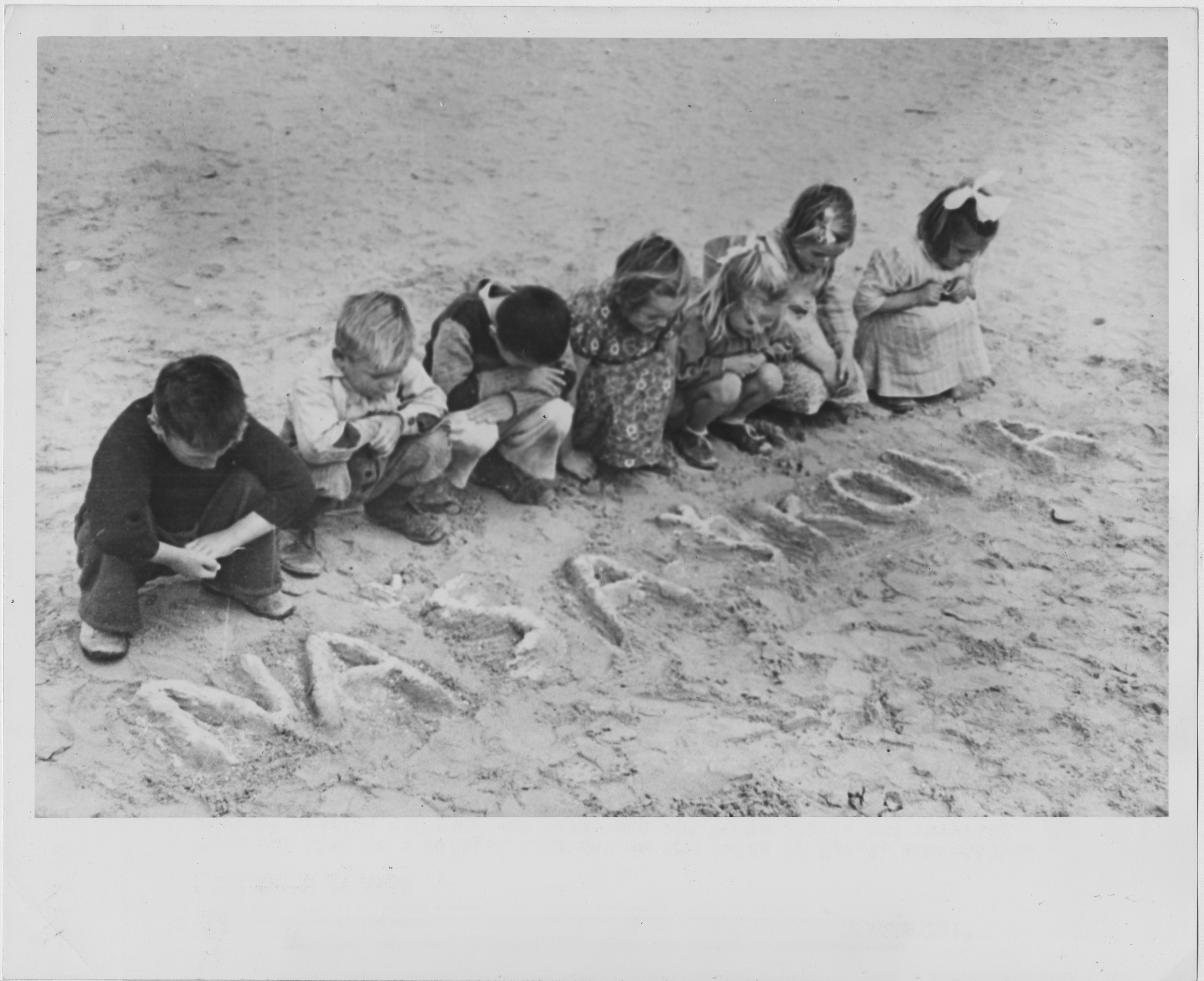 A line of seven young children squat down in sand. They are not looking at the camera, but at letters moulded into the sand in front of them: NAŠA ŠKOLA [our school]